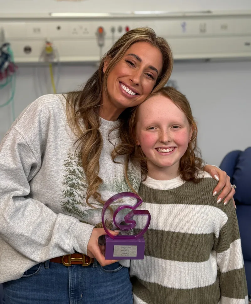 Kirsty Waugh, in a striped jumper, smiling beside Stacey Solomon in a hospital room, holding a purple JustGiving award trophy