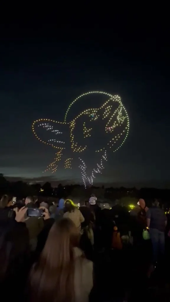 A wolf howling at a full moon, drawn in white and gold drone lights above the crowd