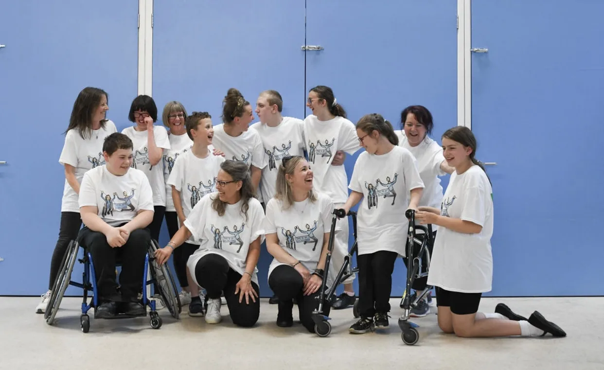 About fifteen This Is Us dancers and helpers in matching white T-shirts posing together in a sports hall, some kneeling at the front and two dancers in wheelchairs