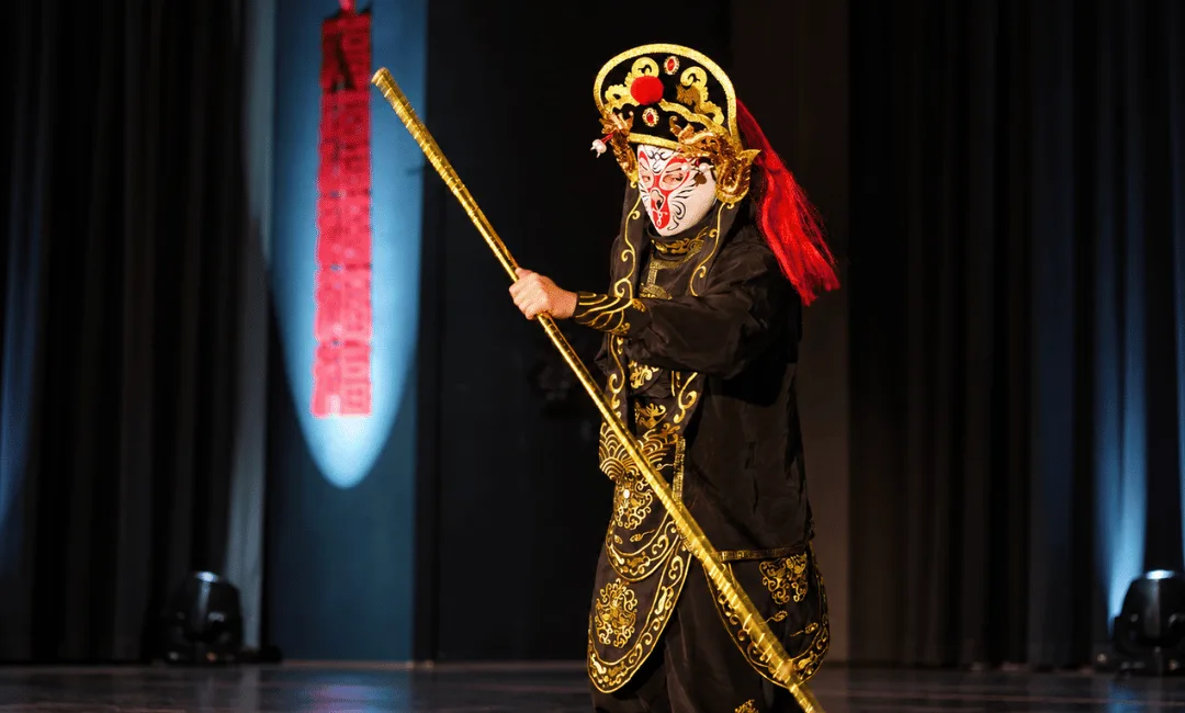 A face-changing performer in black and gold Sichuan Opera costume and a painted mask, holding a golden staff on a dark stage